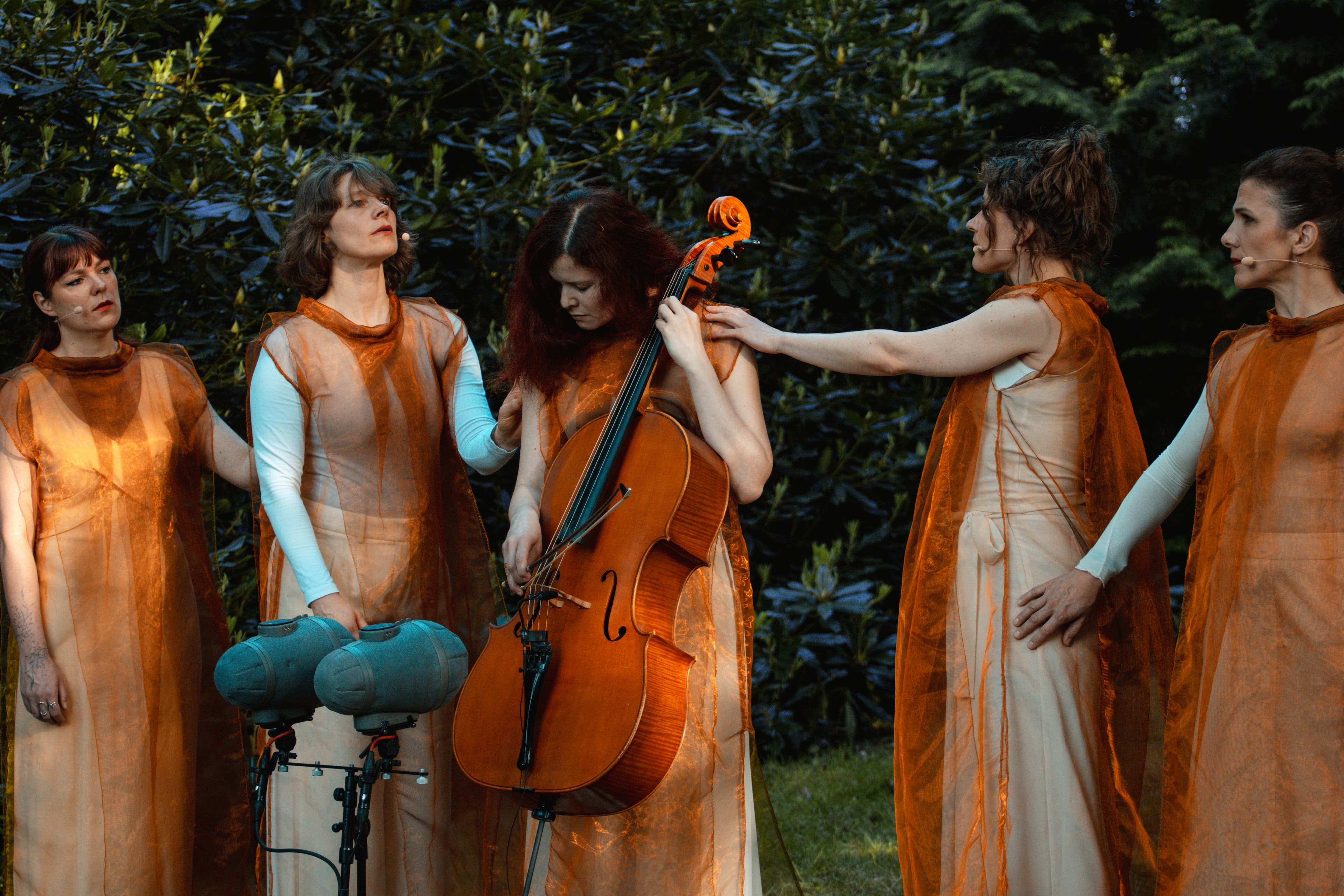 Maya stands with her eyes closed, holding a cello in her hands, surrounded by trees, with two singers on either side of her.