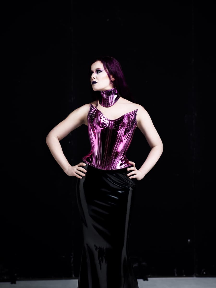 Maya Fridman standing hands on hips in a metallic pink corset and choker, black skirt, against a black backdrop