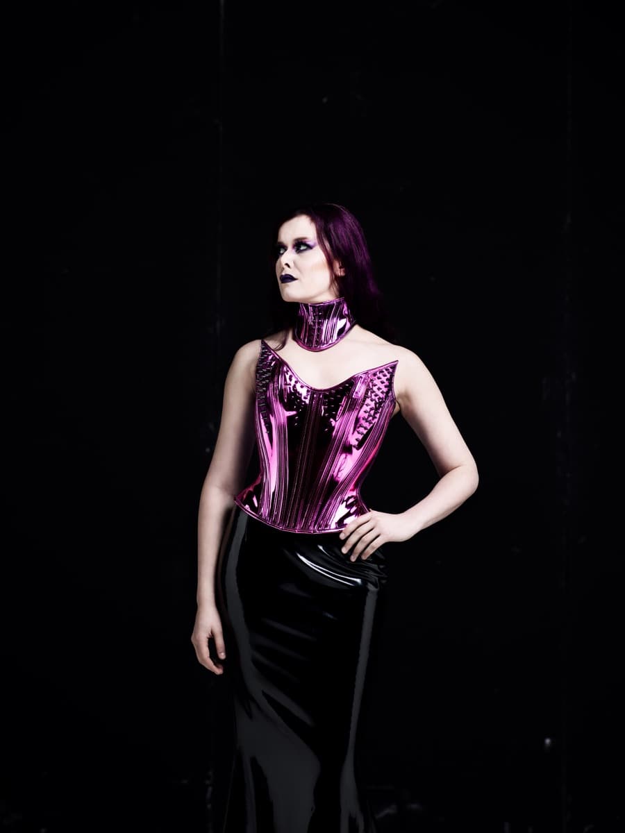 Maya Fridman in a metallic pink corset and choker, one hand on her hip, gaze lifted, against a black backdrop