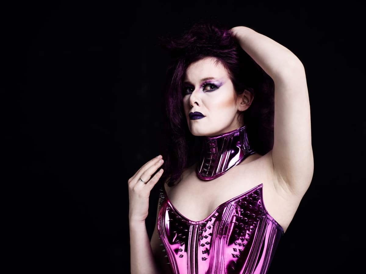 Maya Fridman, close portrait, purple hair held up with one hand, metallic pink corset against a black backdrop